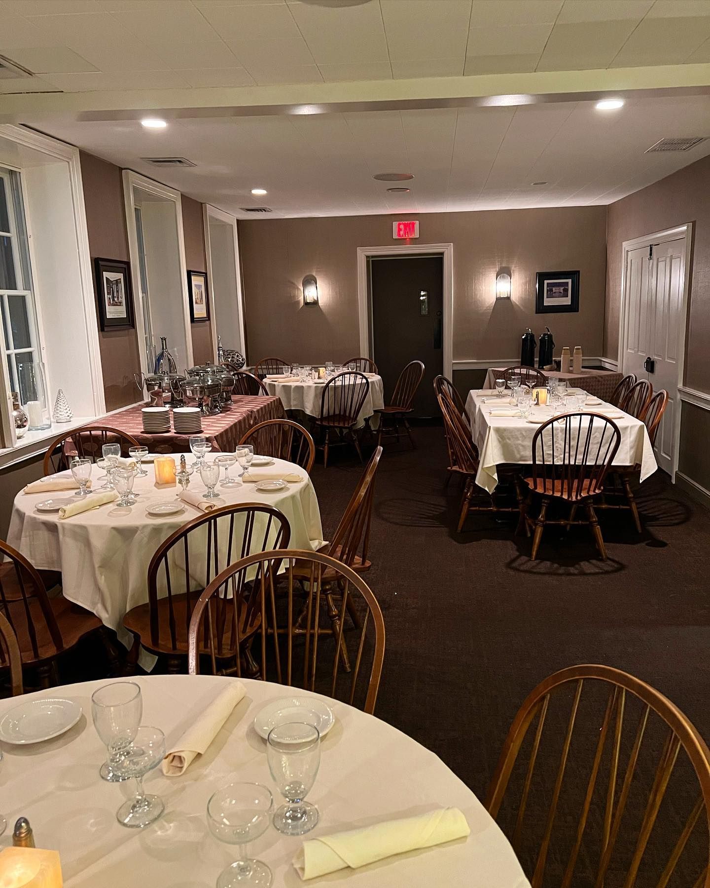 Empty banquet room with round tables set for dining and a lit exit sign at the back.