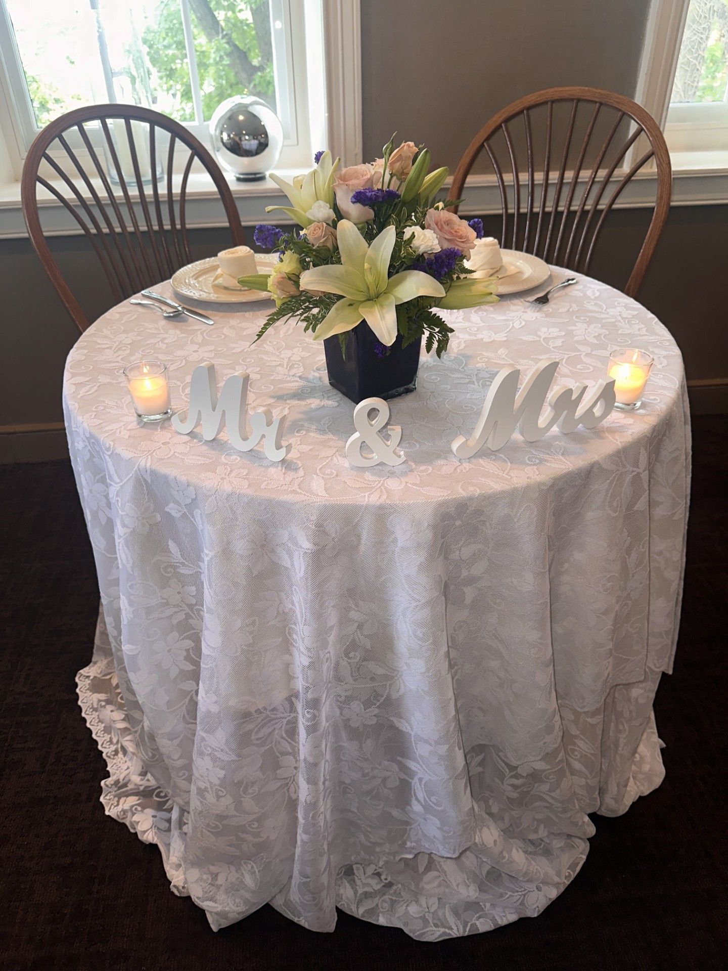Decorated round table with white lace cloth, floral centerpiece, candles, and “Mr & Mrs” sign near a window