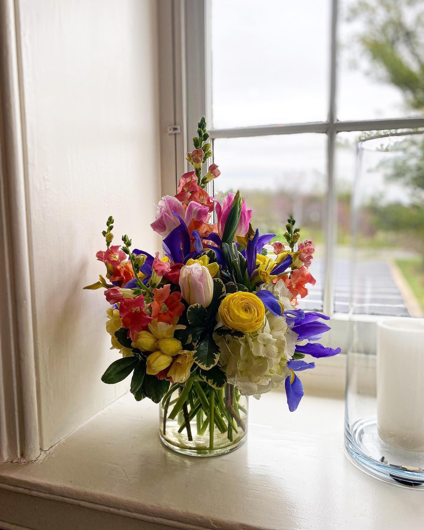 Colorful bouquet of mixed flowers in a glass vase on a windowsill beside a candle holder