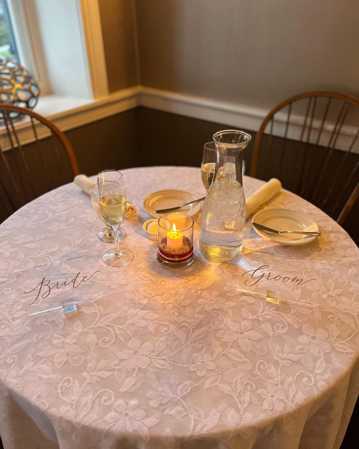 Romantic candlelit table with white tablecloth, glass pitcher, plates, and folded napkins by a window