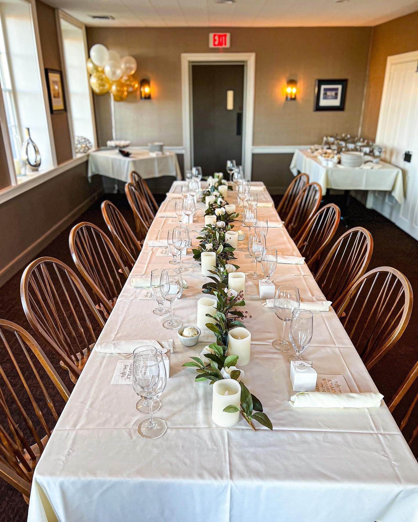Long banquet table set with white linens and floral greenery in a warm indoor dining room