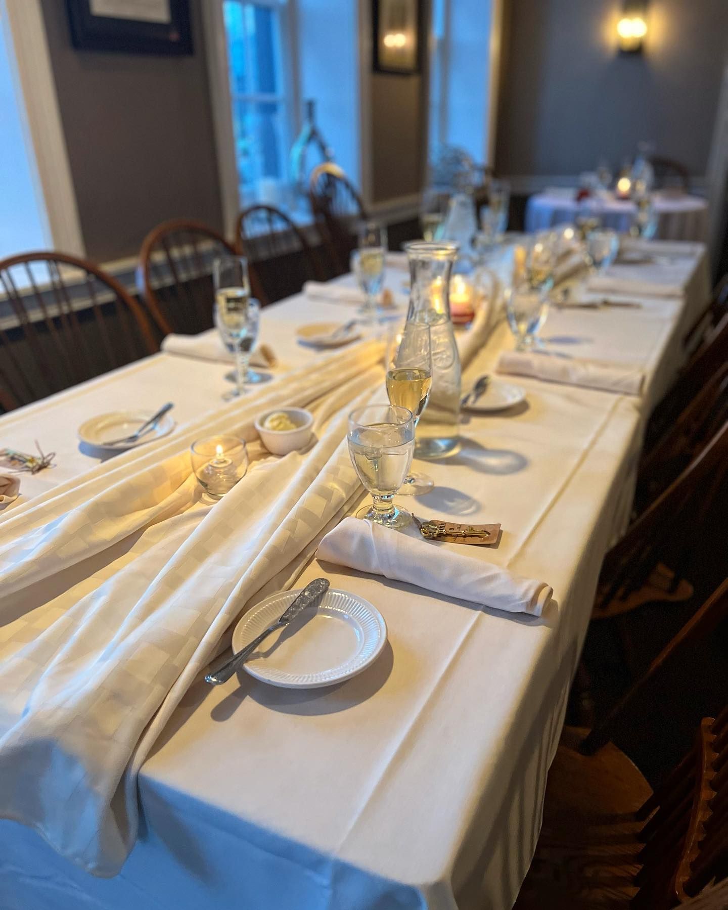 Elegantly set long dining table with white linens, glassware, and folded napkins in a dimly lit restaurant.
