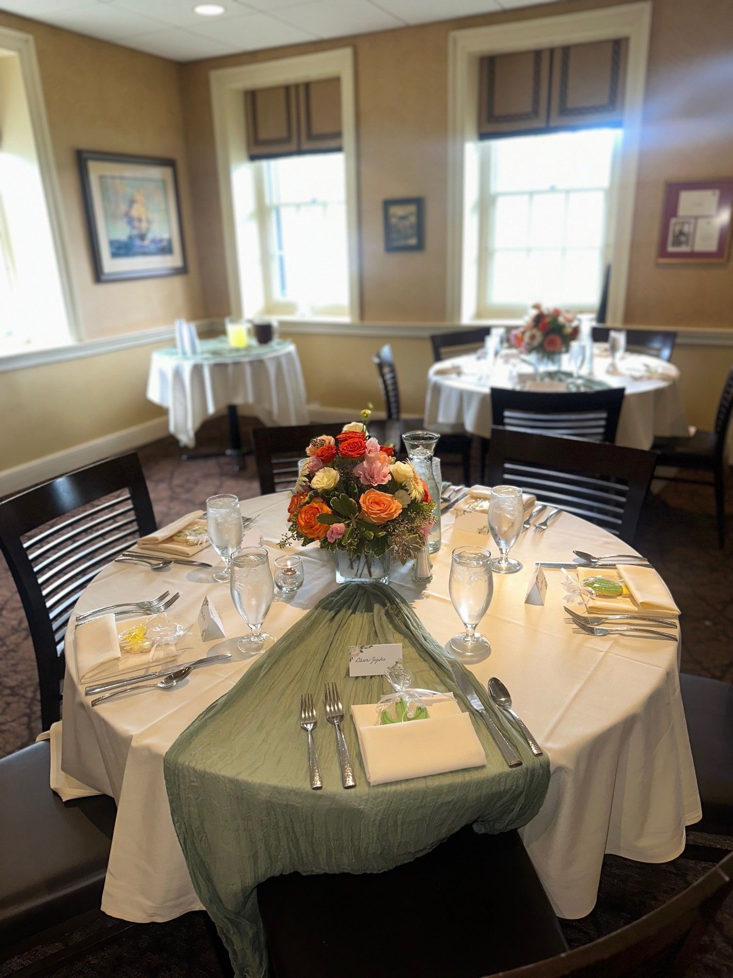 Round banquet table set with white linens, green napkins, place settings, and orange floral centerpiece in a dining room