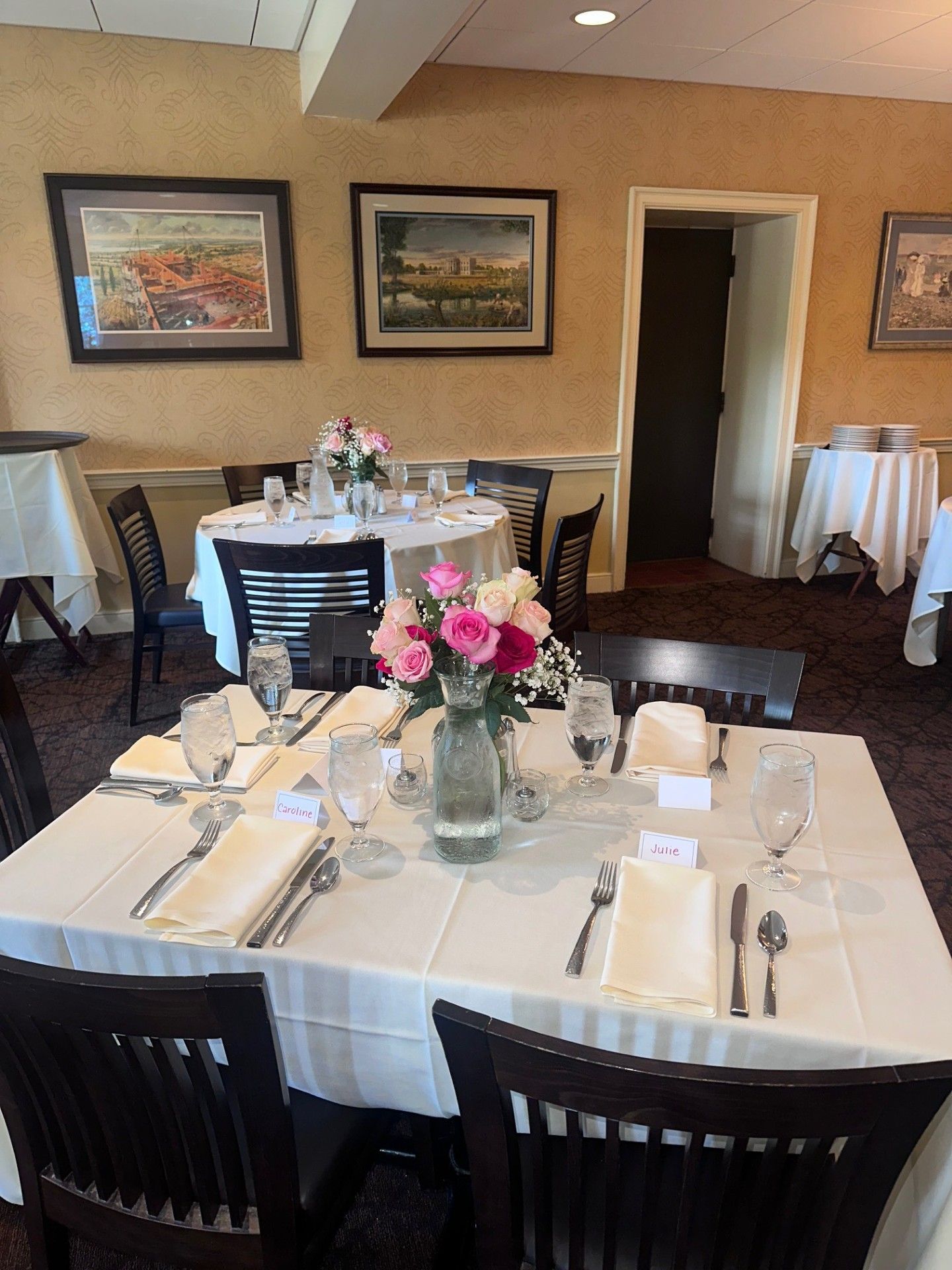 Dining room with set tables, white linens, pink floral centerpieces, and framed wall art.