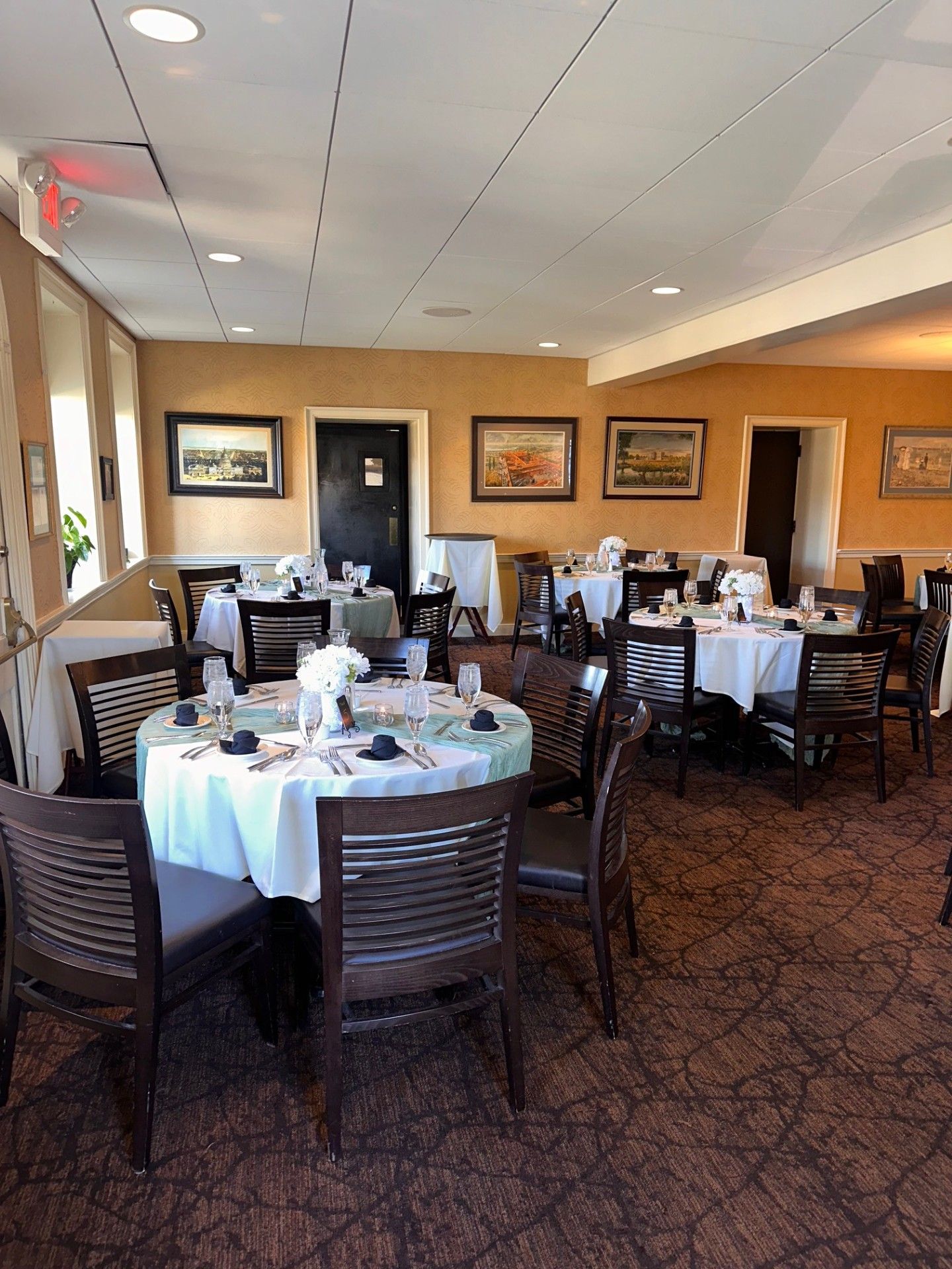 Empty restaurant dining room with set tables, dark chairs, framed wall art, and patterned carpet.