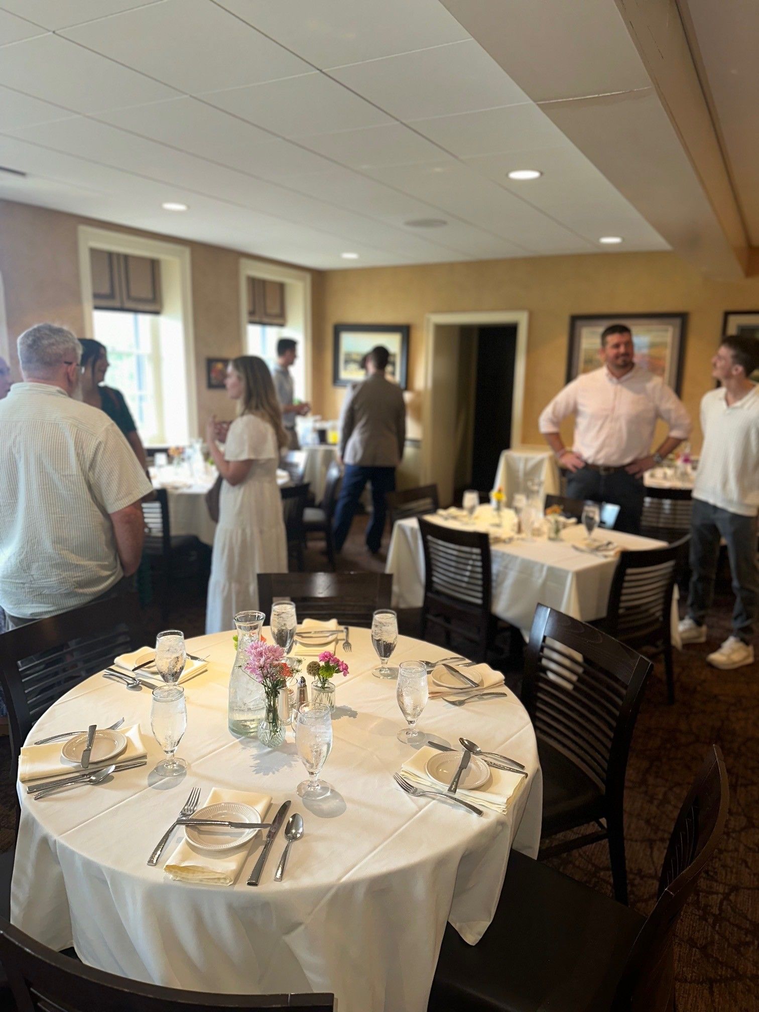 People mingling in a bright banquet room with round tables set for a formal event