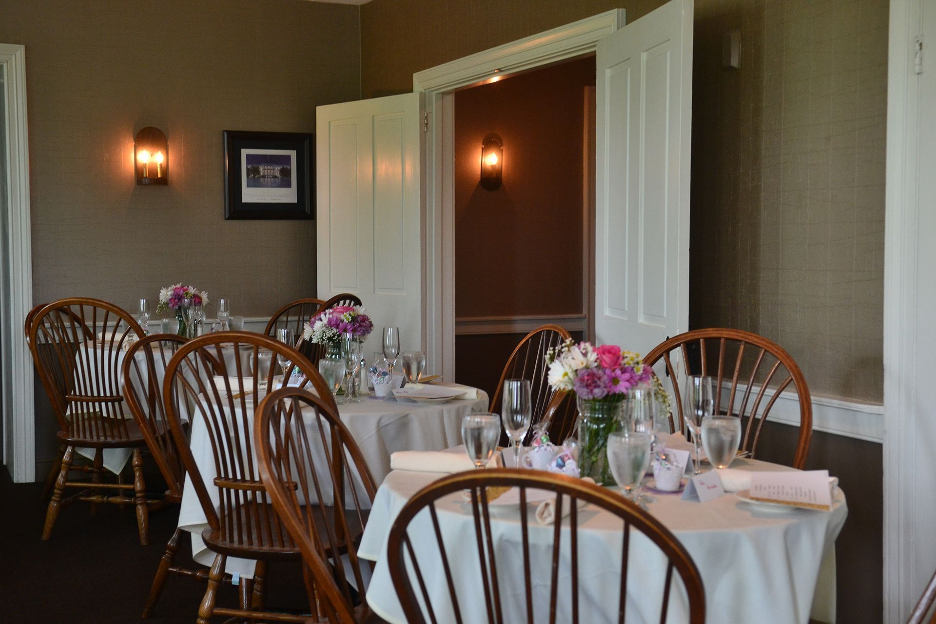 Dining room with set tables, white tablecloths, flowers, and open double doors to a warmly lit adjoining room