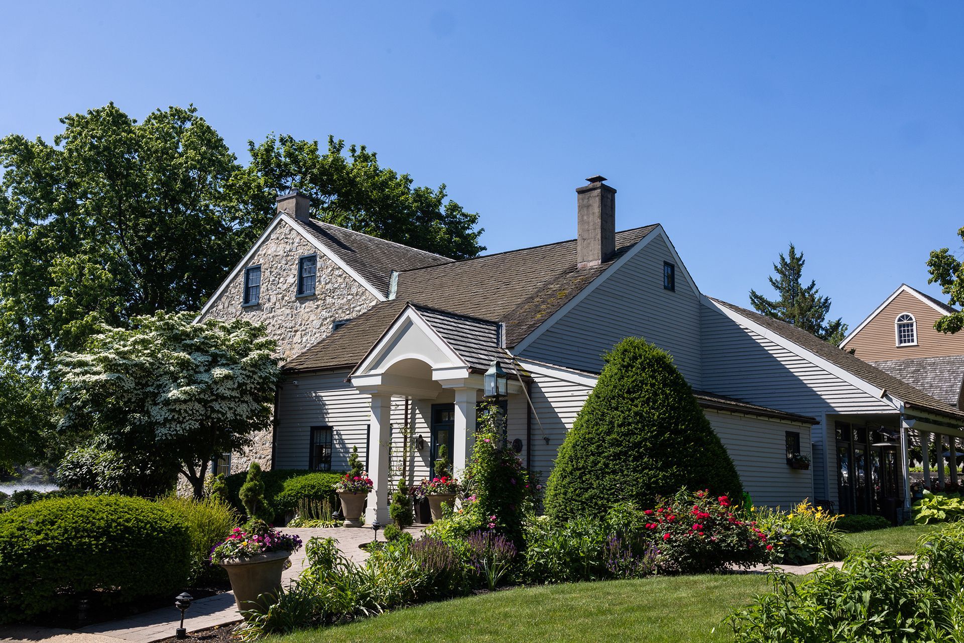 White house with a porch, manicured garden, and trees under a clear blue sky