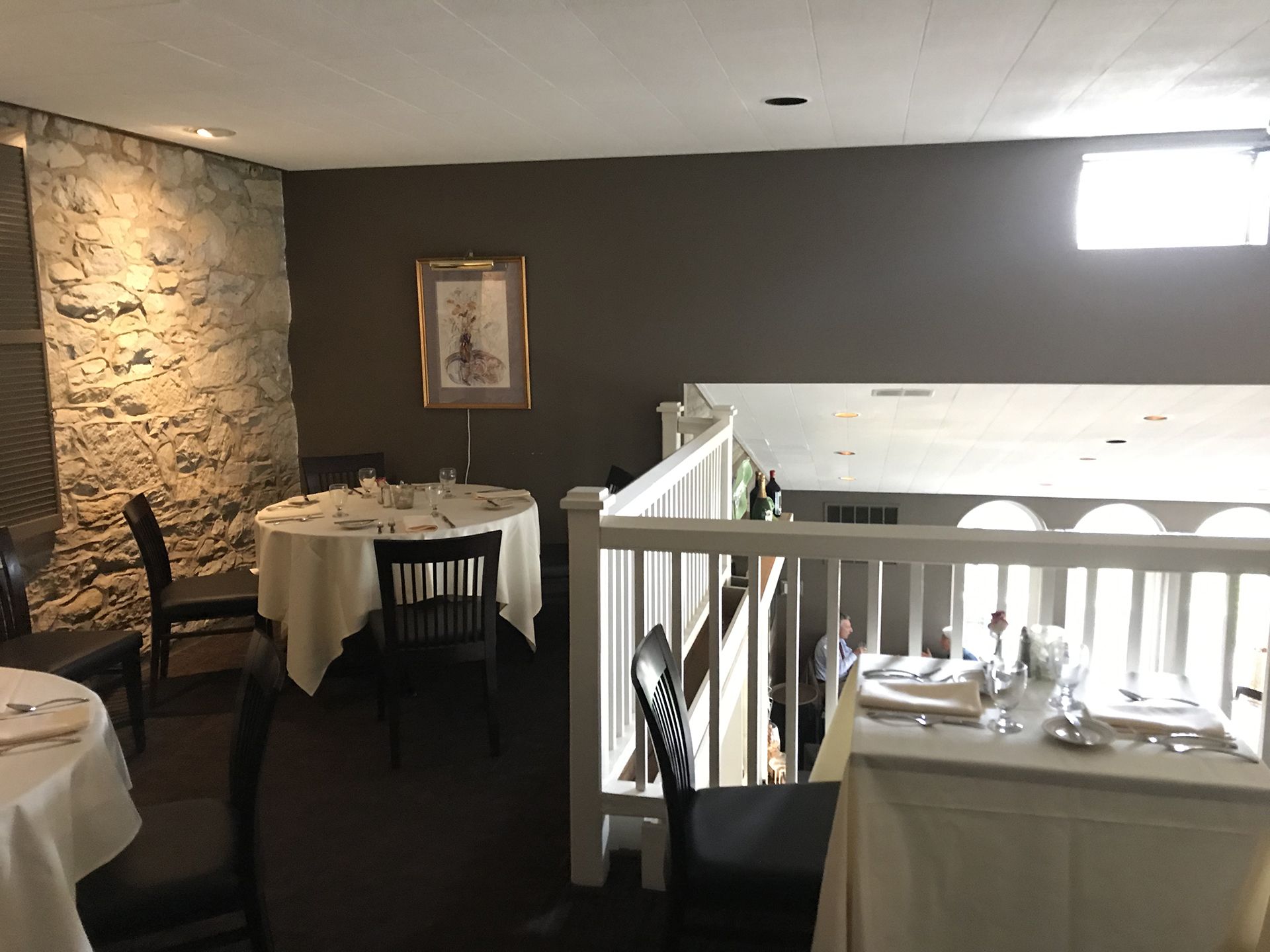 Small dining area with round tables, white tablecloths, dark chairs, stone wall, and balcony railing.