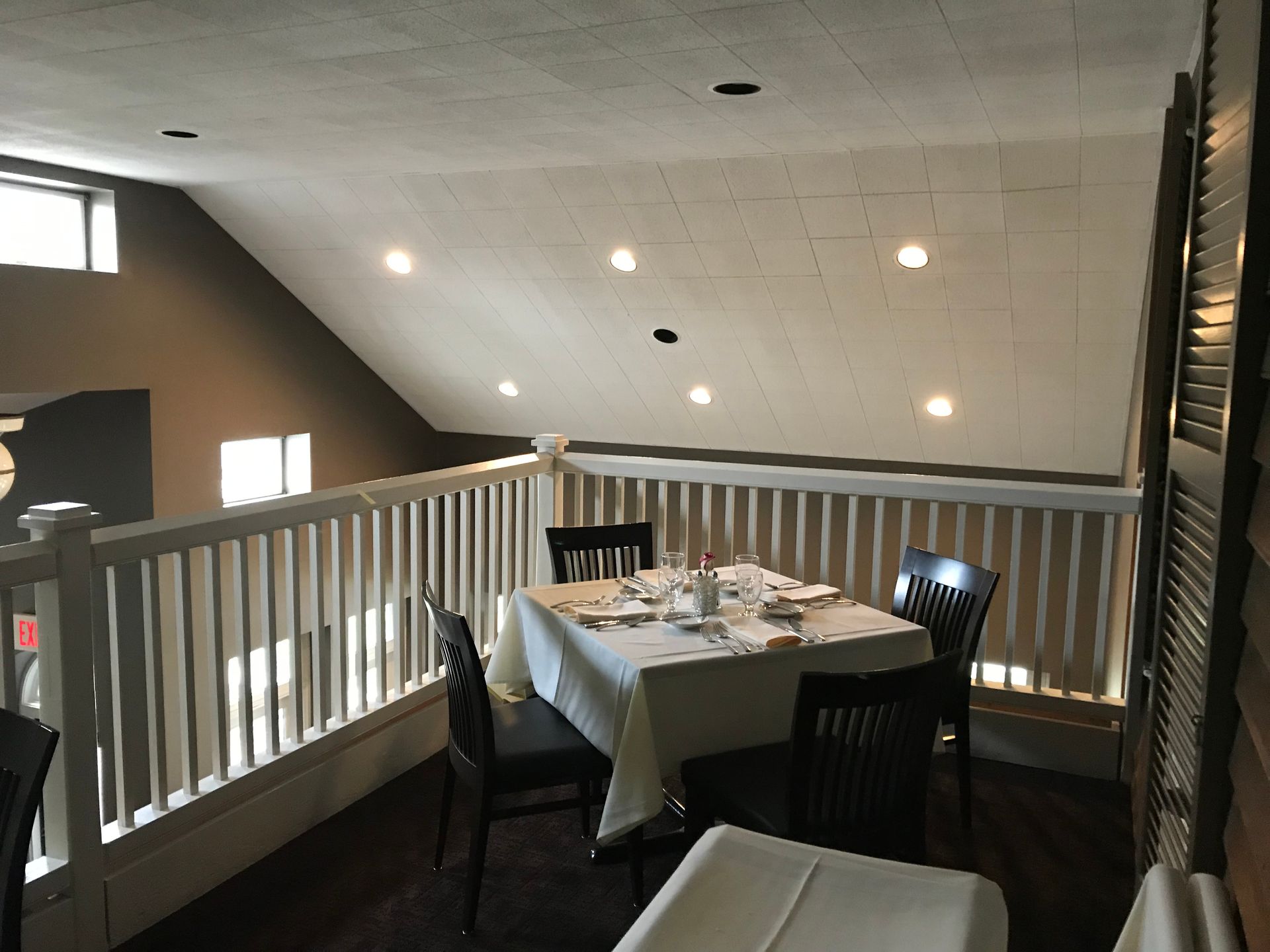 Small dining table with white tablecloth and four chairs on an upstairs balcony overlooking a bright room.