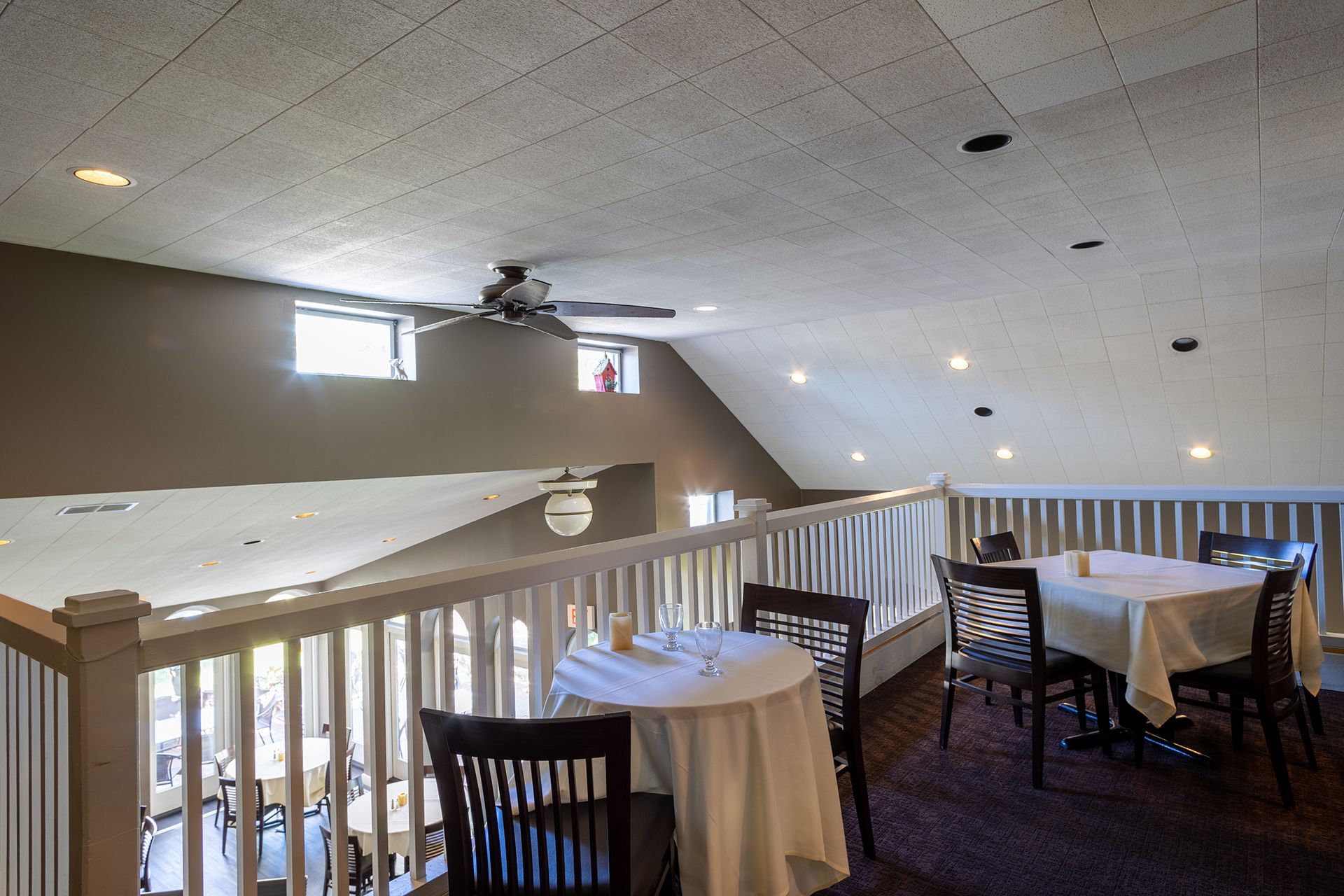 Empty banquet hall with round tables, white tablecloths, and wooden chairs under a sloped ceiling.