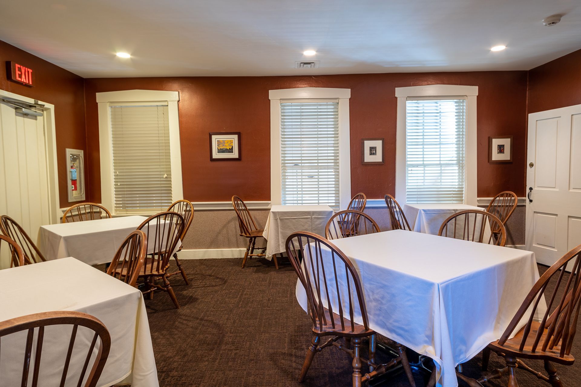 Empty dining room with white-clothed tables, wooden chairs, and windows with blinds