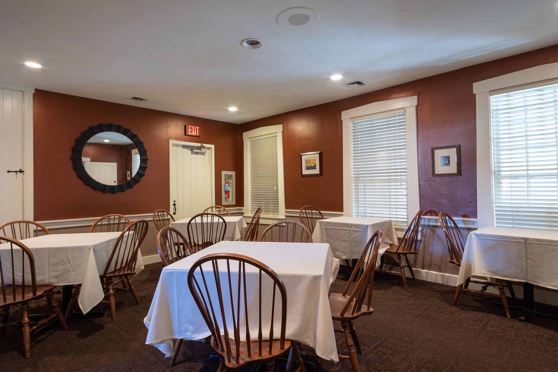 Empty dining room with white tablecloths, wicker chairs, and warm red walls with windows and framed art