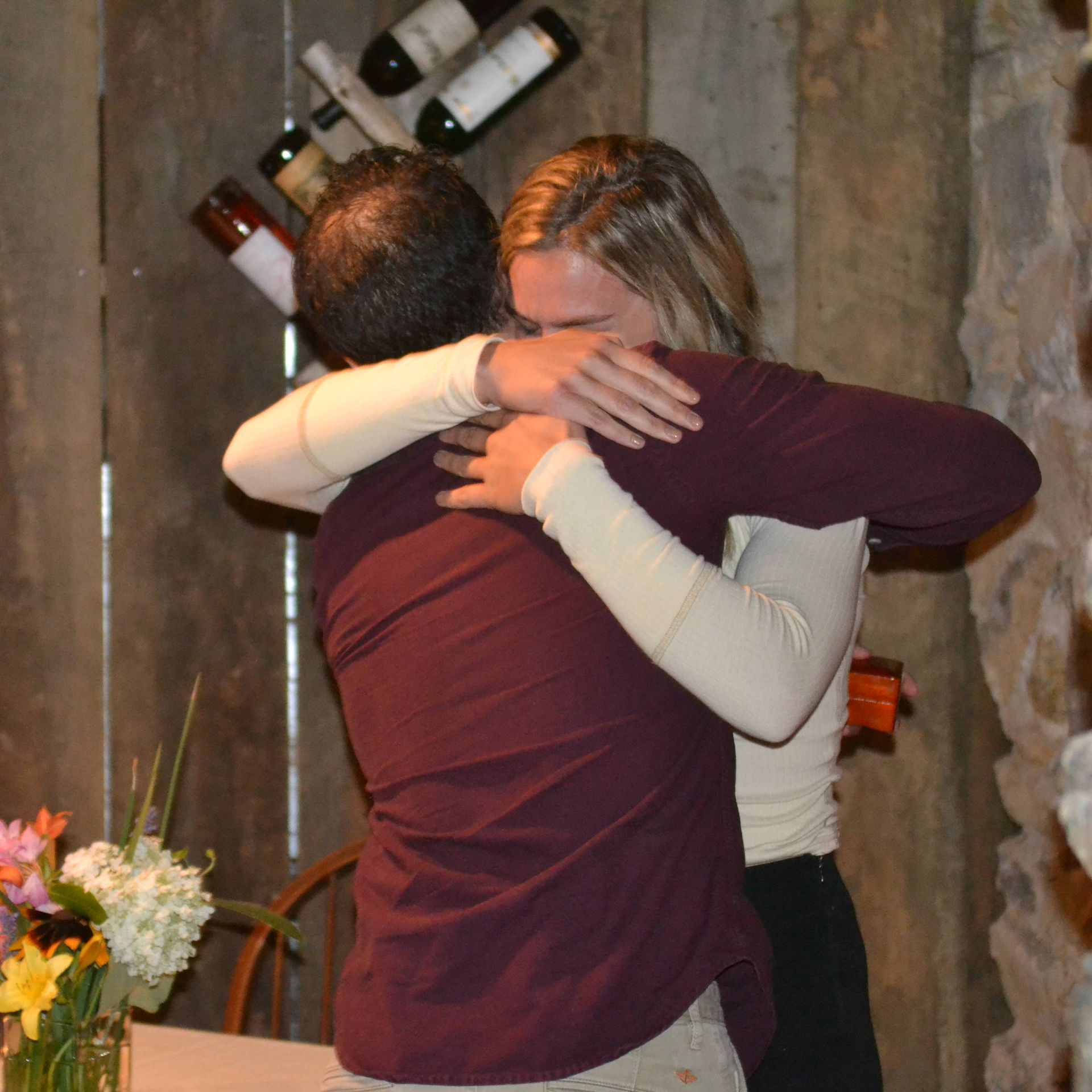 Two people hugging in a rustic room with wine bottles on the wall