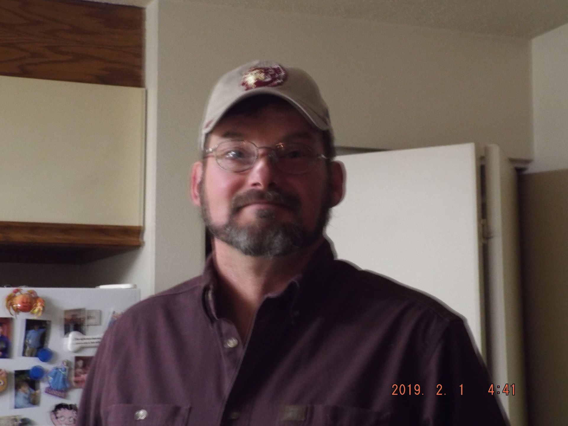 Man with glasses, beard, and cap in a kitchen, wearing a dark button-down shirt.