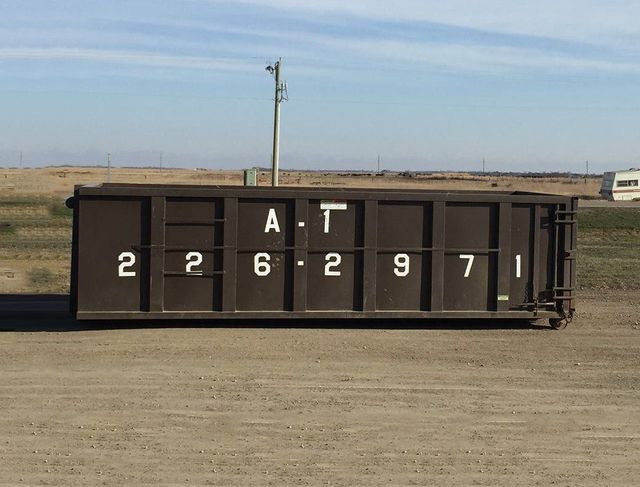 Dark brown dumpster labeled "A-T 2262971" on a gravel surface, with a plain landscape background.
