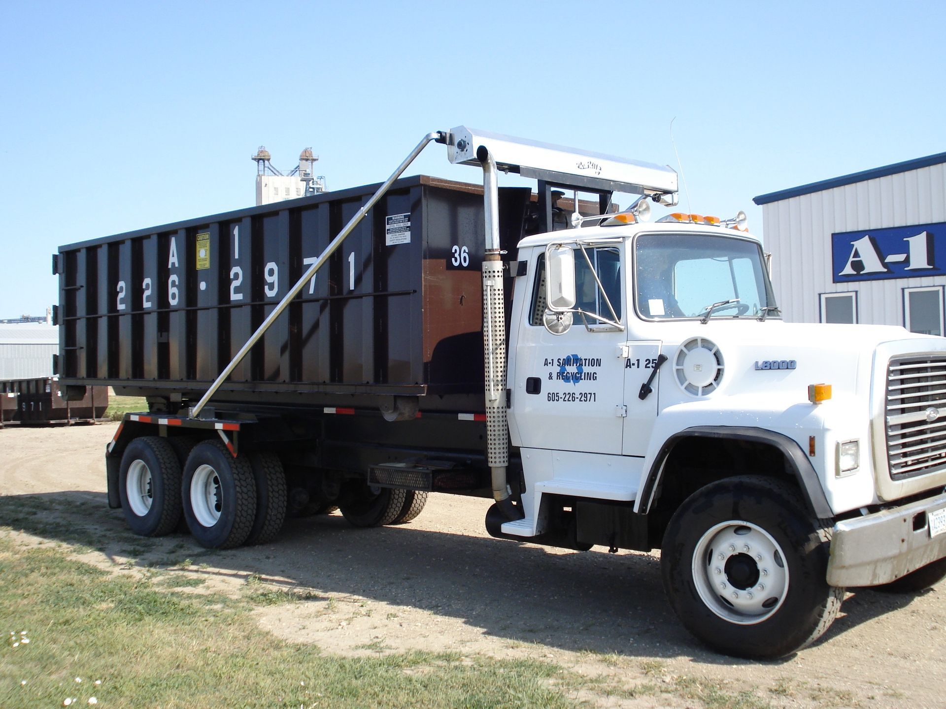 White roll-off truck with a black container in front of a building on a sunny day.