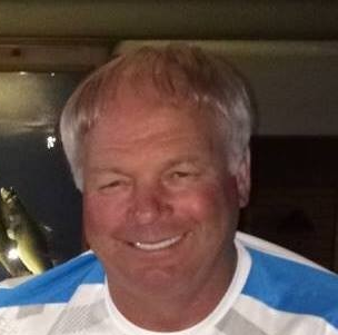 Man with fair hair smiling in front of a blurry background. He is wearing a blue and white shirt.