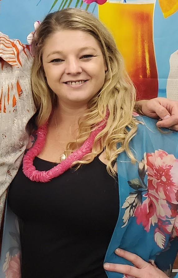 Woman smiles, wears pink lei, black top, floral blue shawl, blonde hair. Luau-themed backdrop.
