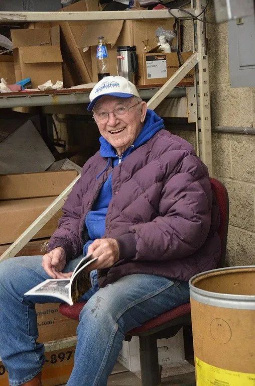 A man in a purple jacket is sitting in a chair reading a magazine