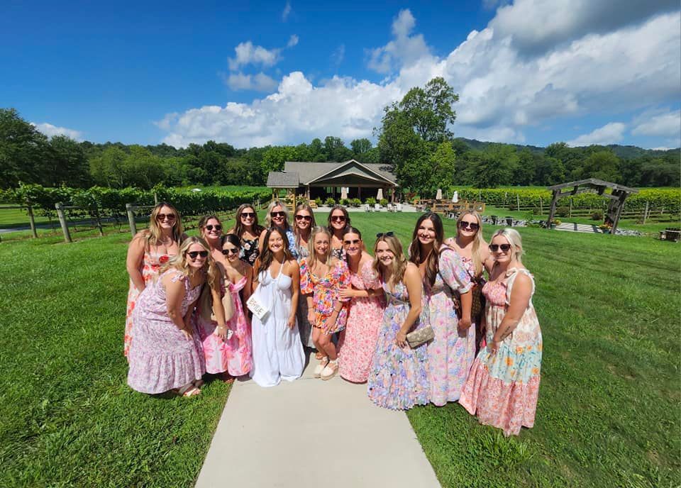 A group of people in floral dresses posing on a lawn in front of a rustic building and vineyard under a sunny blue sky.
