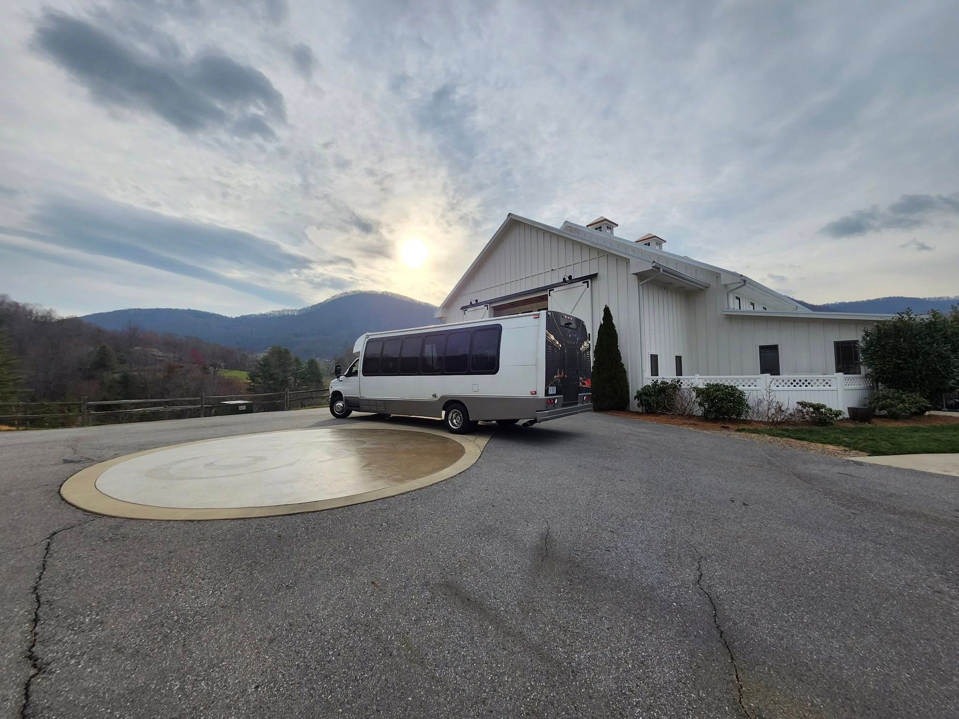 A white passenger bus parked on an asphalt driveway in front of a white barn-style building with mountains in the distance.