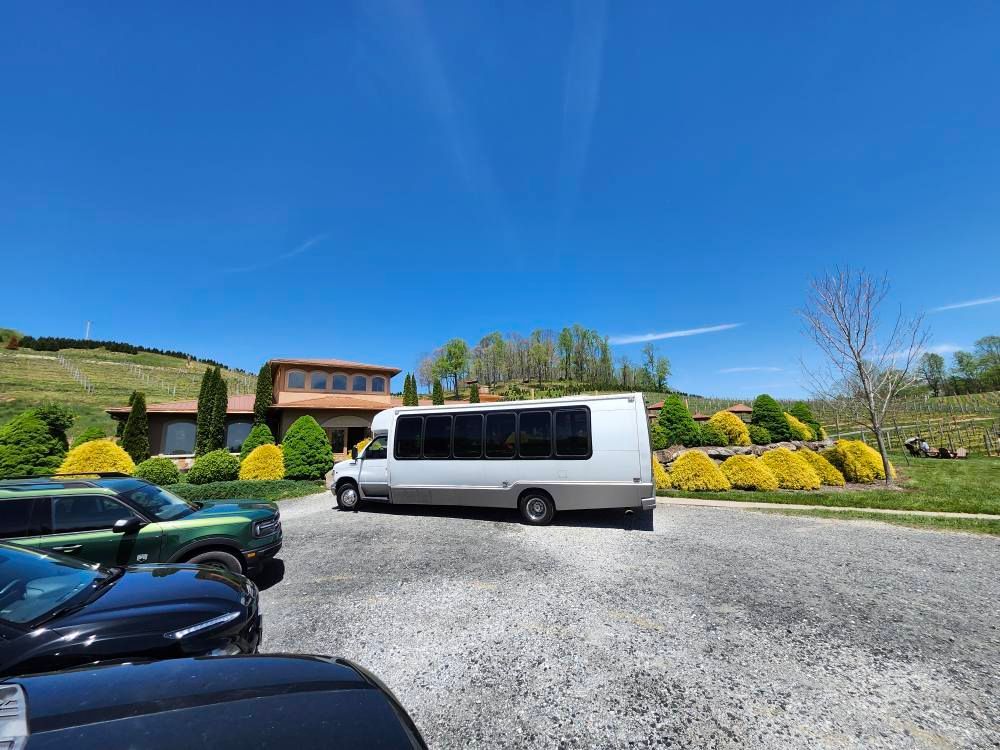A white passenger shuttle bus parked on a gravel lot in front of a building with rolling green hills under a blue sky.