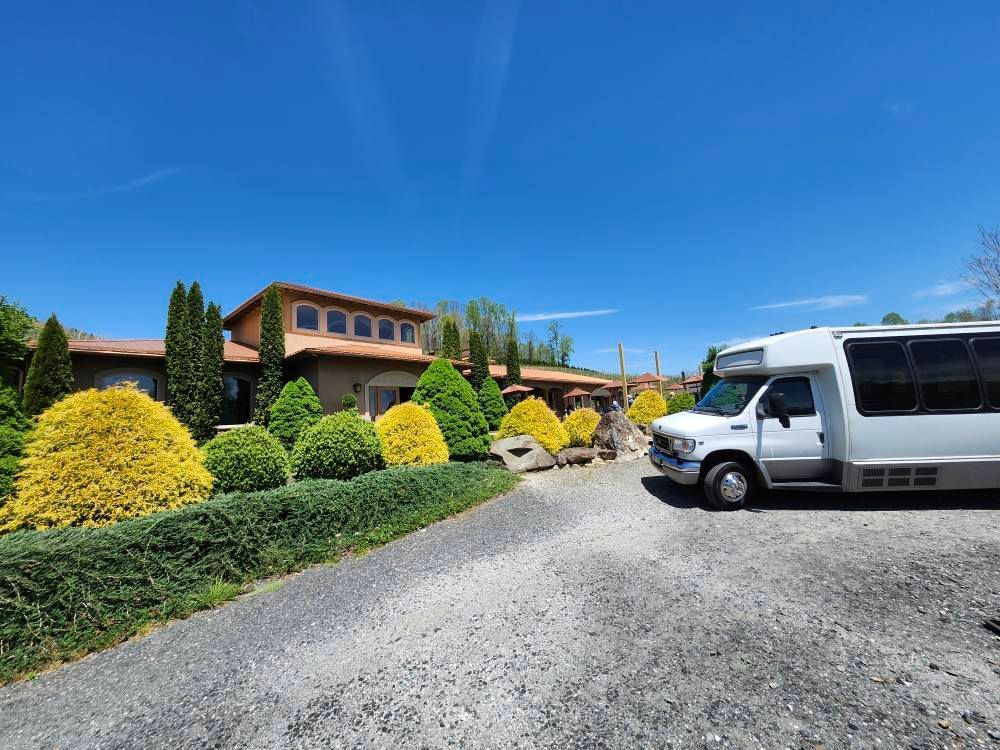 A white shuttle bus parked on a gravel driveway in front of a tan house with a terracotta roof under a clear blue sky.