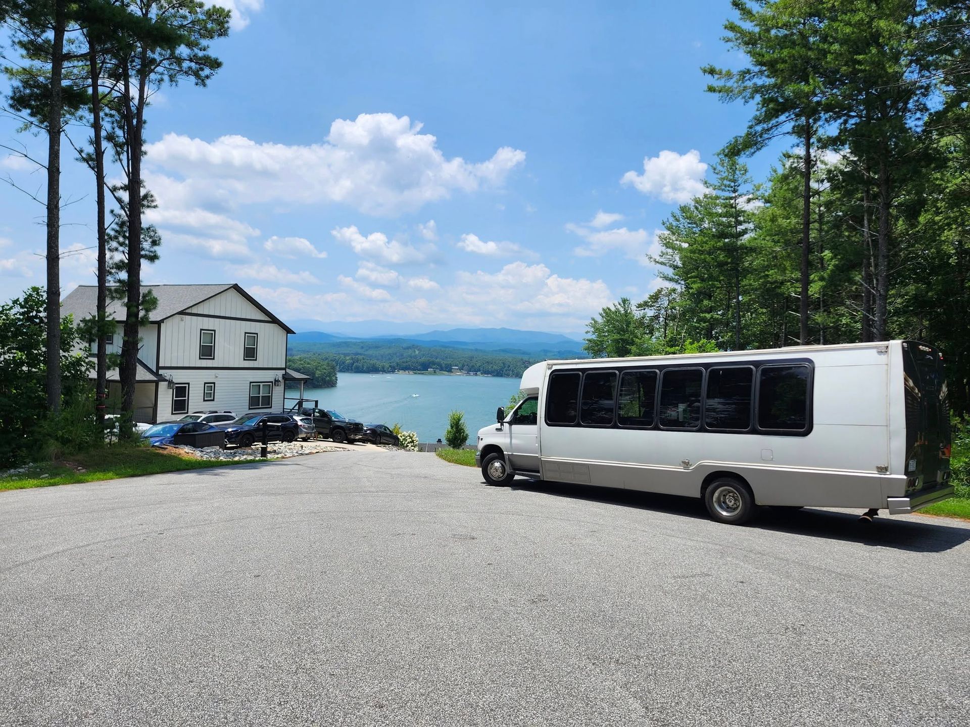 A white shuttle bus parked on a gravel lot overlooking a lake and distant mountains next to a white house.