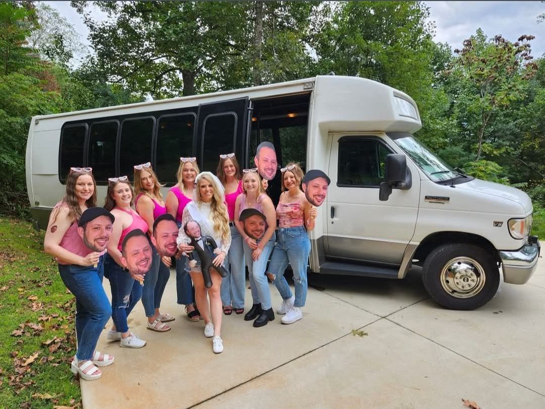 A group of people wearing pink and holding large face cutouts stand before a white shuttle bus in a wooded setting.