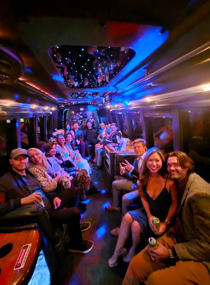 A group of people sitting inside a party bus with blue ceiling lights and a starlit mirror, smiling towards the camera.