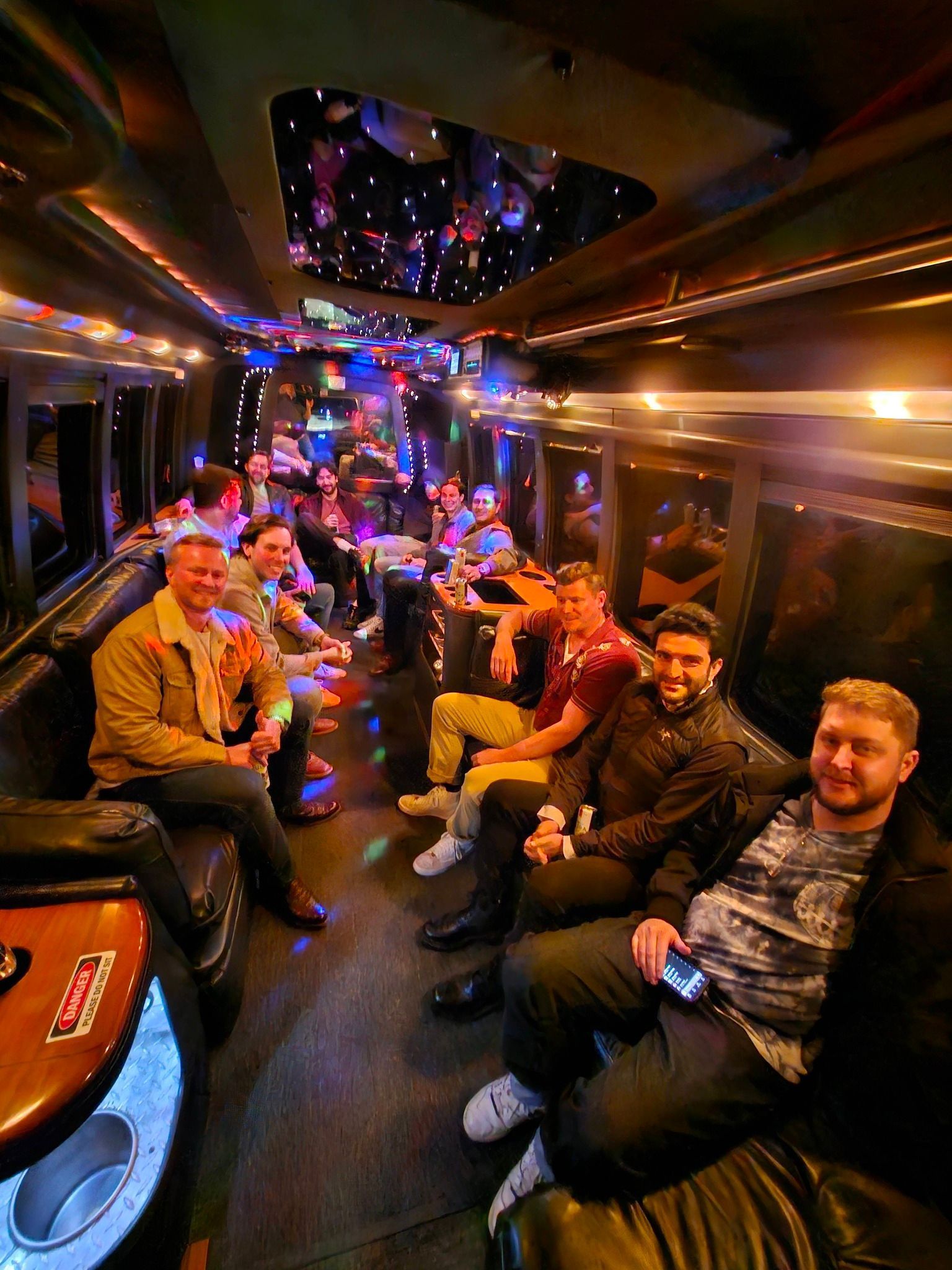 A group of people sitting inside a party bus with colorful ambient lighting, facing the camera.