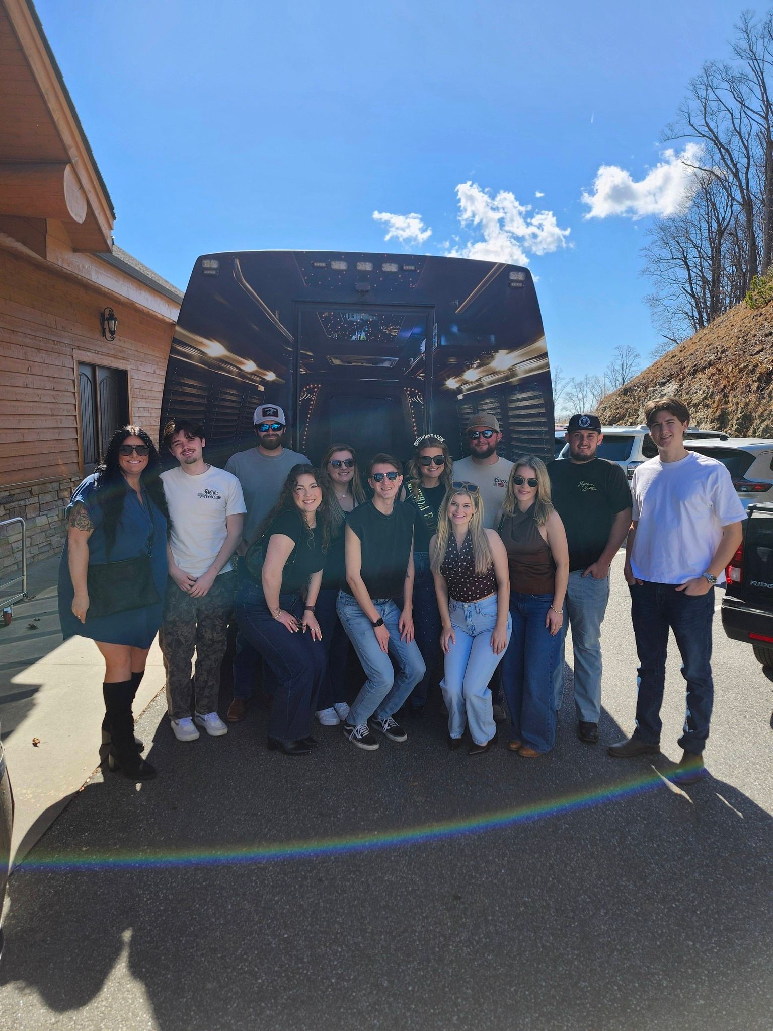 A group of people posing for a photo in front of a dark, patterned shuttle bus on a sunny day.