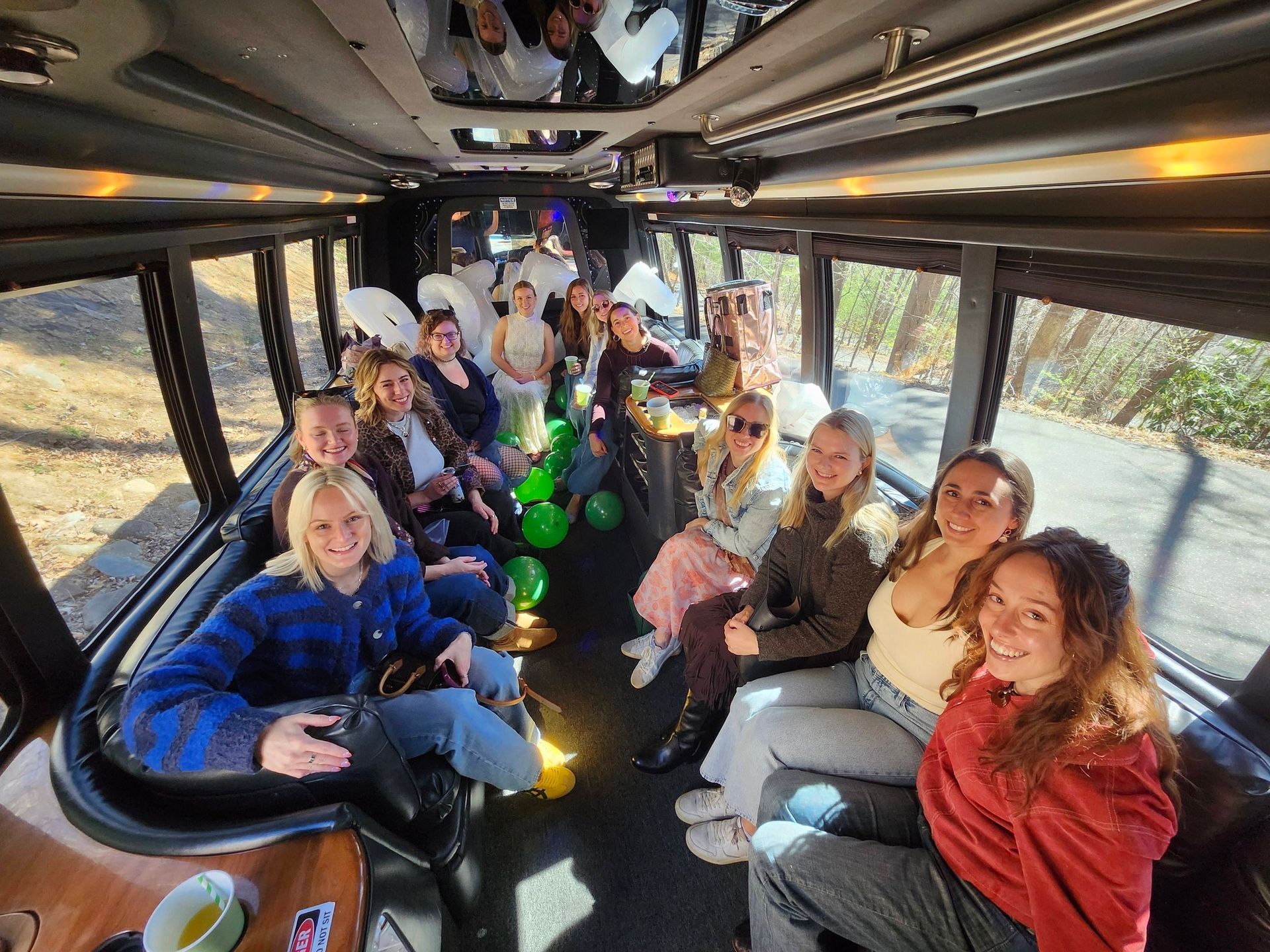 A group of people sitting inside a party bus filled with green balloons, looking toward the camera and smiling.