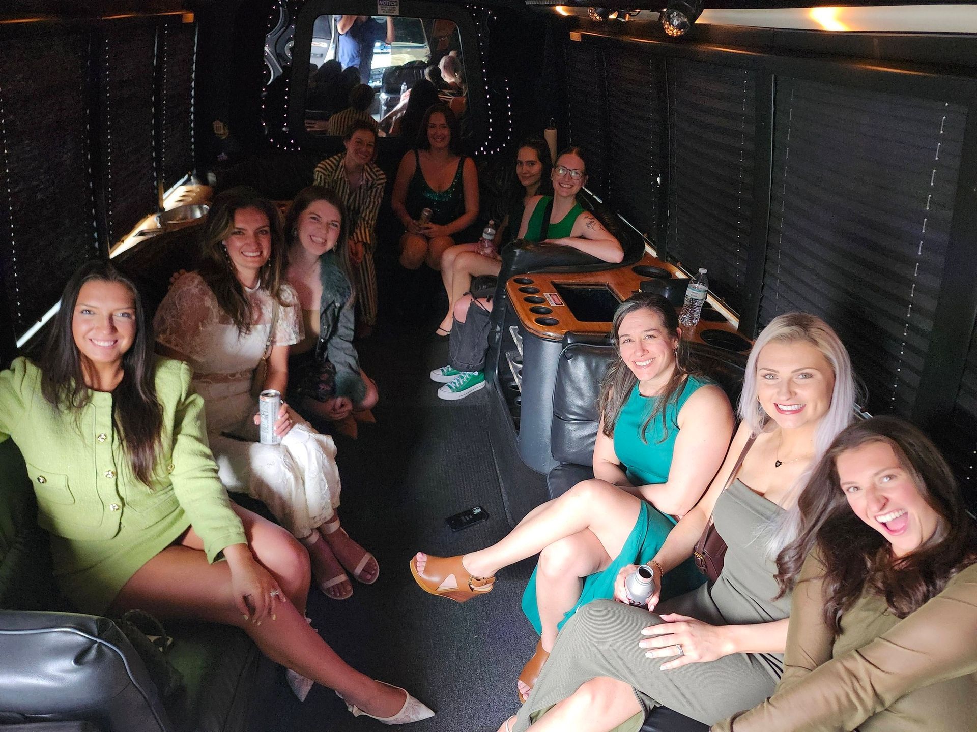 A group of women in formal attire socializing and laughing inside a dimly lit party bus with black leather seating.