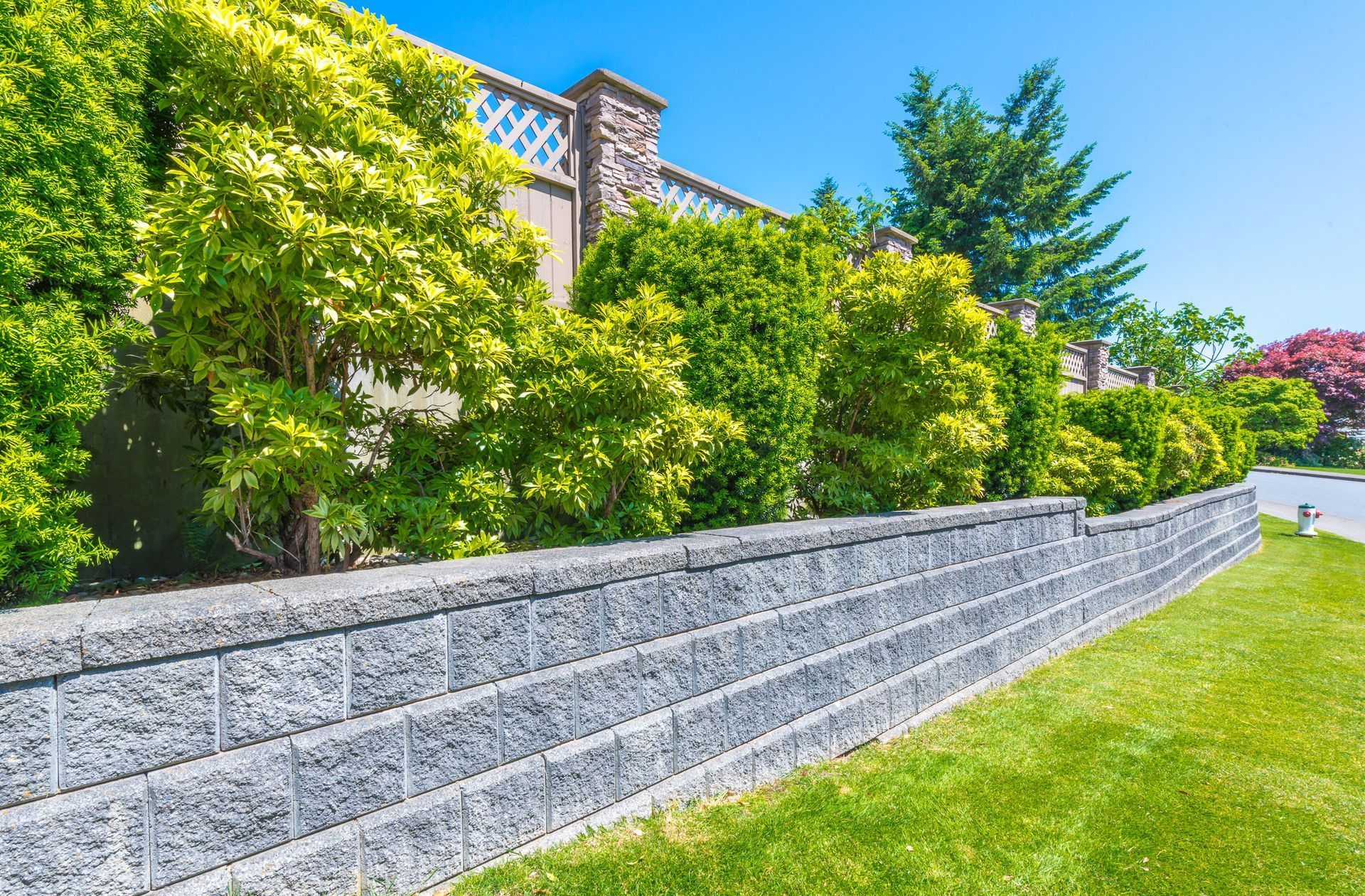 A gray stone retaining wall borders a green lawn with lush green trees and a decorative stone fence in the background.
