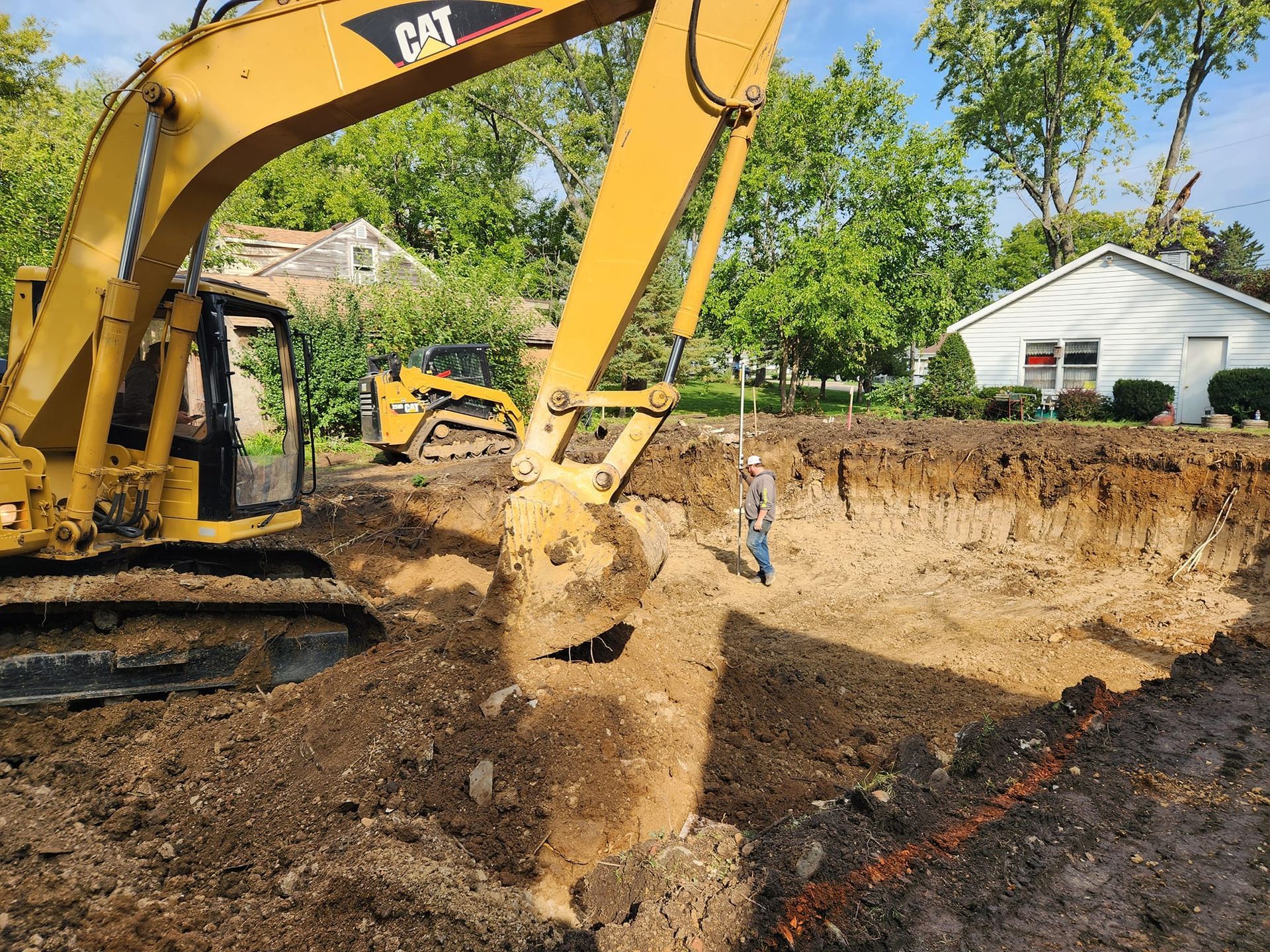 A yellow CAT excavator digs a large hole in a residential yard while a construction worker stands in the pit nearby.
