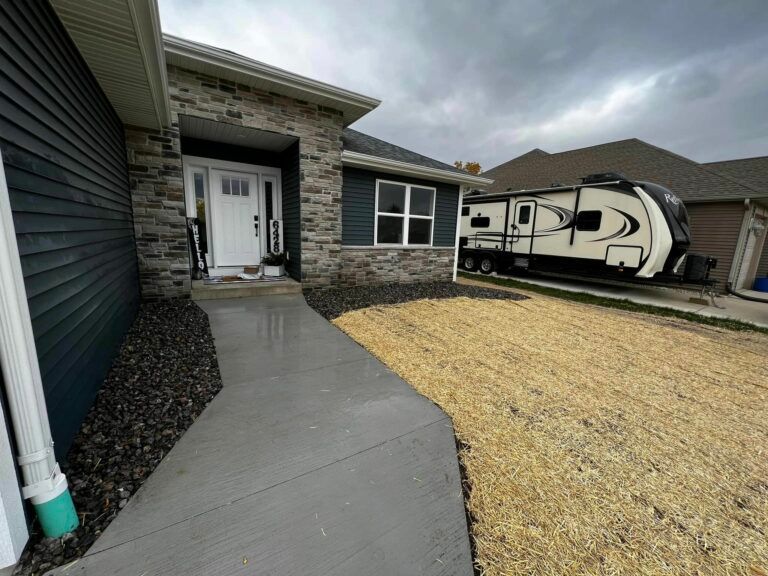 A front entrance featuring a white door, grey stone siding, a concrete walkway, and a large RV parked on the side.