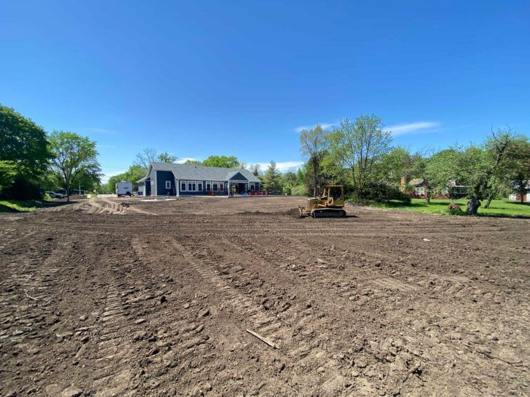 A yellow construction bulldozer sits on a large, freshly graded dirt lot in front of a modern grey house.