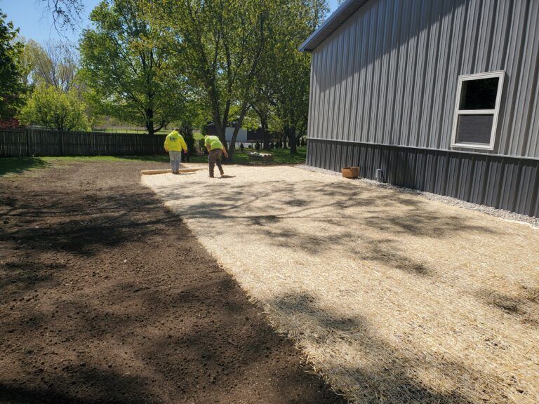Two workers in high-visibility vests work on a yard with areas of dark soil and light-colored straw mulch near a building.