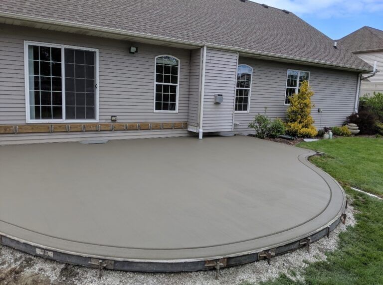 A newly poured, curved concrete patio against the back of a light gray vinyl-sided house with a grassy yard.
