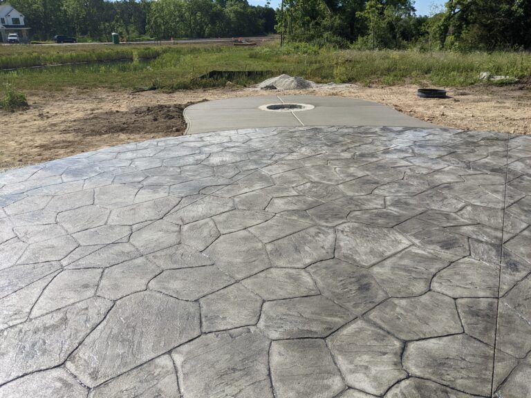 A grey, stamped concrete patio with a random stone pattern leads to a smooth, circular concrete slab in a grassy backyard.