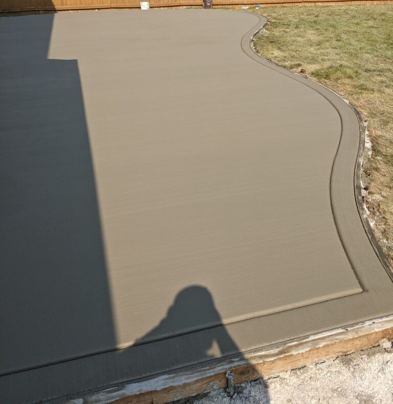 A freshly poured, smooth concrete patio with a curved decorative border edge, situated next to a grassy yard.