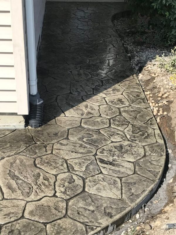 Stamped concrete walkway with a stone-pattern design and grey coloring, located beside a house exterior.