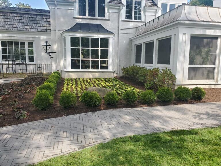 A white multistory home with a manicured garden featuring spherical boxwood shrubs and a herringbone brick walkway.