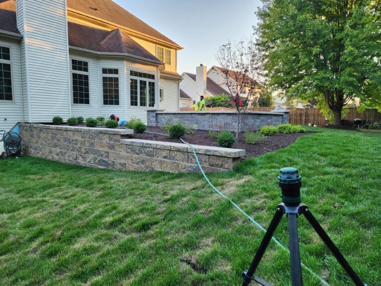 A view of a tan multi-level house with a stone retaining wall in the backyard and a tripod with equipment in the grass.