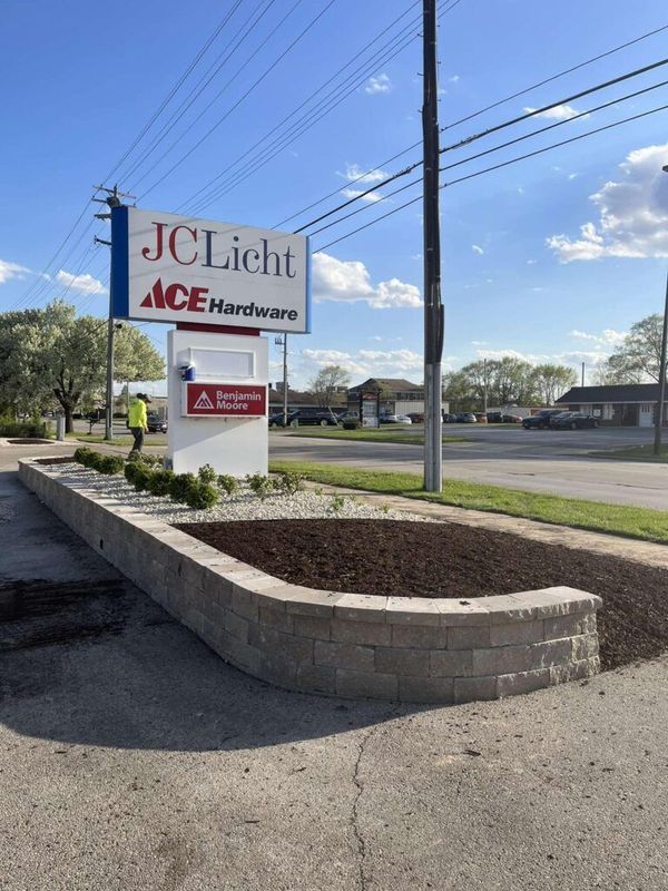 A JC Licht and Ace Hardware sign stands above a freshly mulched landscaped area with a concrete retaining wall.