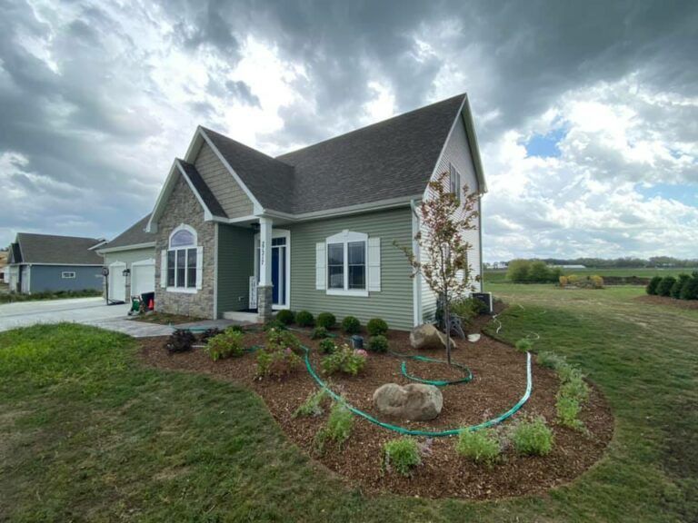 A two-story green house with stone accents, a multi-gabled roof, and a landscaped front yard under a cloudy sky.