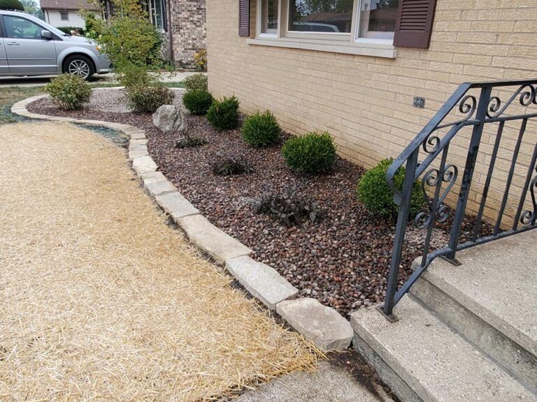 A house foundation bed with dark mulch, several small green shrubs, a stone border, and a black metal handrail.