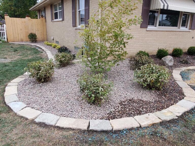 Landscaped garden bed with shrubs and a young tree, bordered by stone blocks in front of a brick house.